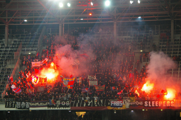 Eine große Menschenmenge in einem Stadion hält Fahnen und Banner, mit Leuchtraketen und Rauch, der aus ihnen aufsteigt, während Metallrahmen und Deckenleuchten über ihnen sichtbar sind.