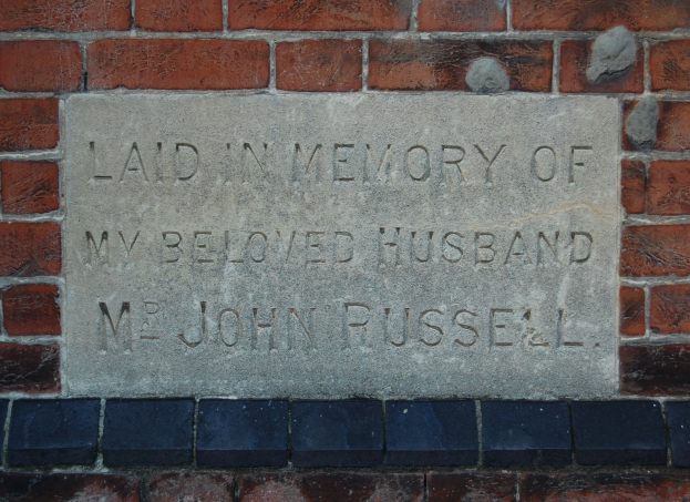 A brick wall with a stone plaque mounted on it, reading "Laid in Memory of My Beloved Husband".