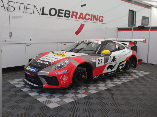 Ein Porsche 911 GT3 RSR in einer Garage, positioniert auf dem Boden mit einer Wand, die Text und eine Tür im Hintergrund zeigt.