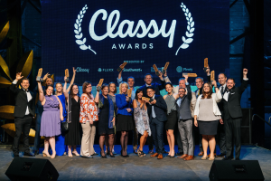 Gruppe von Menschen auf einer Bühne, die Preise halten, mit Rednern darunter und einem Bildschirm im Hintergrund, der "Classy Awards 2019" anzeigt.