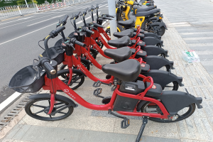 A row of red and black e-bikes parked along a street with a white pole in the foreground, trees and buildings in the background, and a clear blue sky.