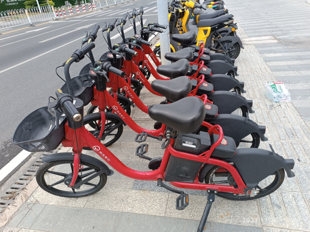 A row of red and black e-bikes parked along a street with a white pole in the foreground, trees and buildings in the background, and a clear blue sky.