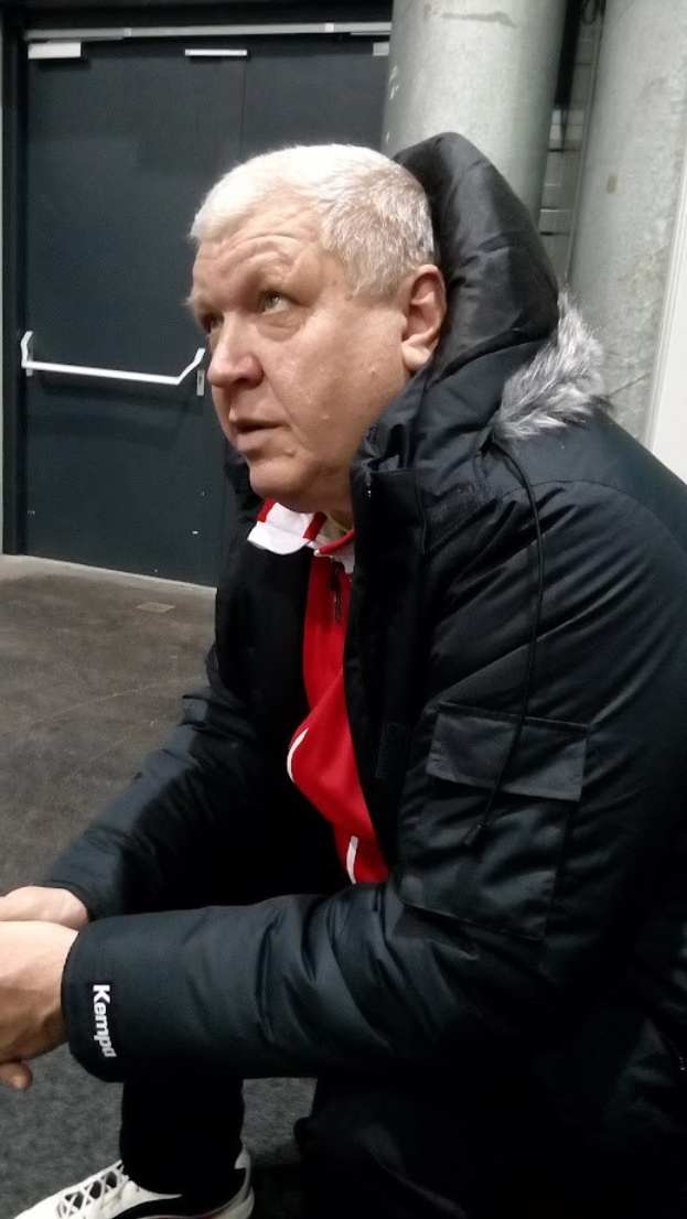 Ein Mann in einer schwarzen Jacke und einem roten Hemd sitzt auf dem Boden vor einer Tür mit einem Griff, einer Säule und einer Wand und scheint tief in Gedanken versunken.