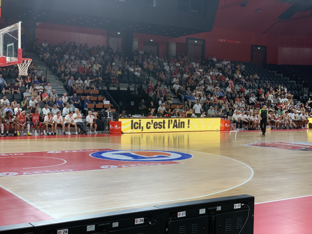 Ein Basketballfeld mit Zuschauern in den Rängen, ein Netz auf der linken Seite und elektronische Geräte unten, in einer Turnhalle mit Treppen, Geländern, Türen, Wänden und Lampen.