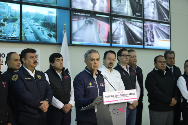 Gruppe von Männern neben einem Rednerpult mit Mikrofon und einer Tafel mit der Aufschrift "Mexikanische Zoll- und Grenzschutzbehörde", vor einer Wand mit Fernsehern und einer Flagge.