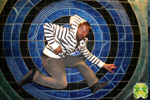 Ein Mann tanzt vor einem bunten Hintergrund mit einem Logo mit Text in der oberen rechten Ecke.