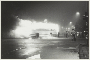 Ein Schwarz-Weiß-Foto eines brennenden Busses am Straßenrand, mit einer Gruppe von Menschen auf dem Gehweg rechts und Straßenlaternen im Hintergrund.