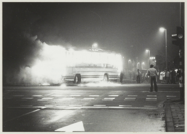 Ein Schwarz-Weiß-Foto eines brennenden Busses am Straßenrand, mit einer Gruppe von Menschen auf dem Gehweg rechts und Straßenlaternen im Hintergrund.