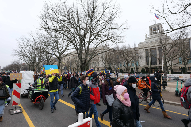 A large group of people participating in a protest march in Washington, D.C. on January 21, 2020, walking down a street with placards, banners, and bicycles, passing buildings and trees under a clear blue sky.