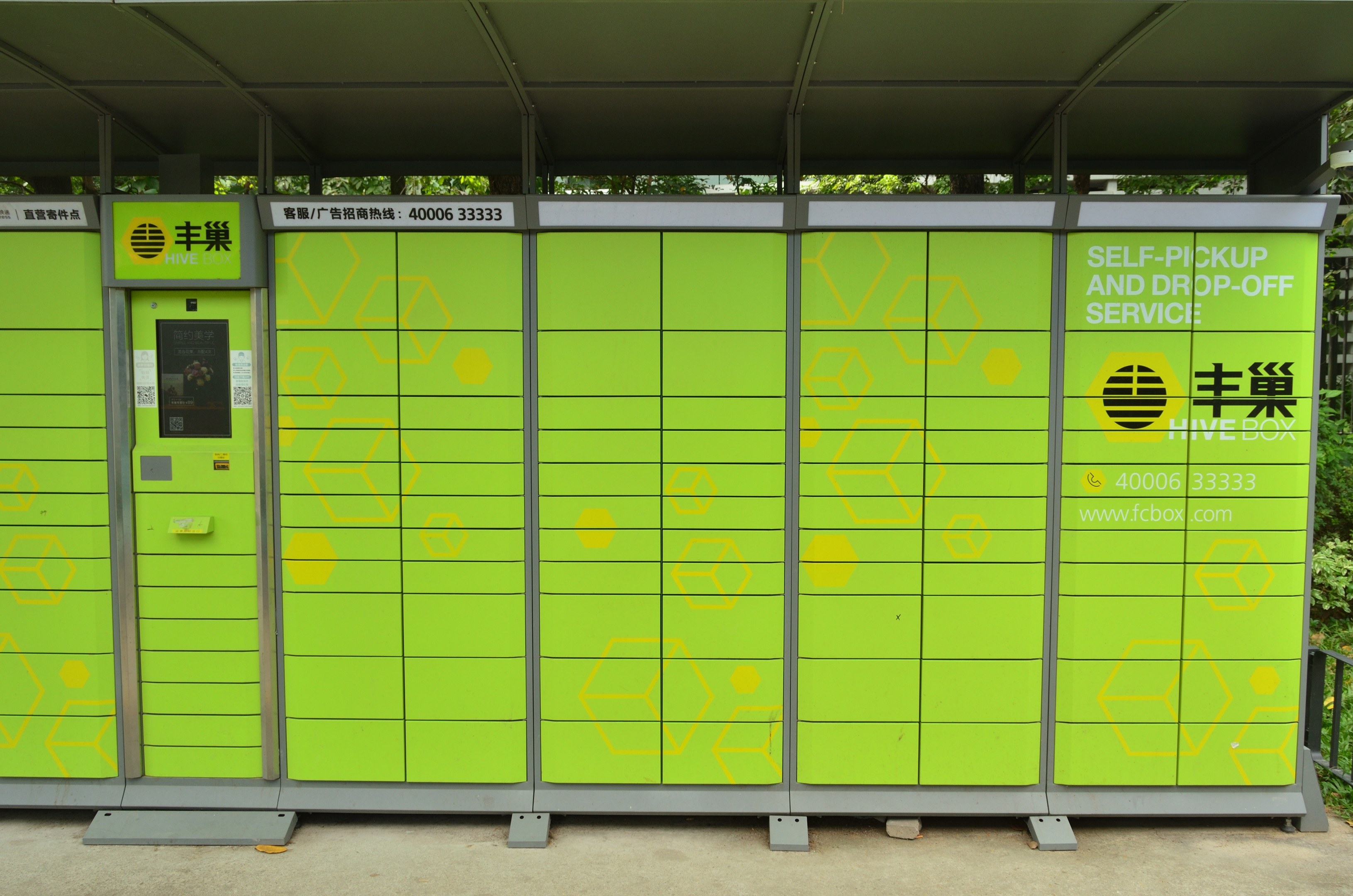 Ein grüner und gelber Verkaufsautomat mit Dach und Text, der neben einer Straße steht, umgeben von einem Metallzaun, Pflanzen, Bäumen und einem bewölktem Himmel, der einen Selbstabhol- und -bringdienst anzeigt.