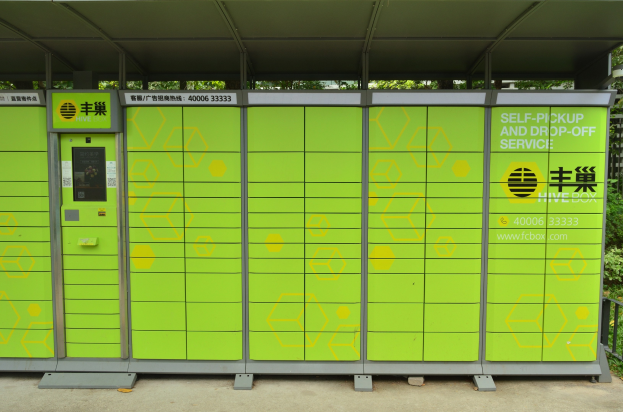 Ein grüner und gelber Verkaufsautomat mit Dach und Text, der neben einer Straße steht, umgeben von einem Metallzaun, Pflanzen, Bäumen und einem bewölktem Himmel, der einen Selbstabhol- und -bringdienst anzeigt.