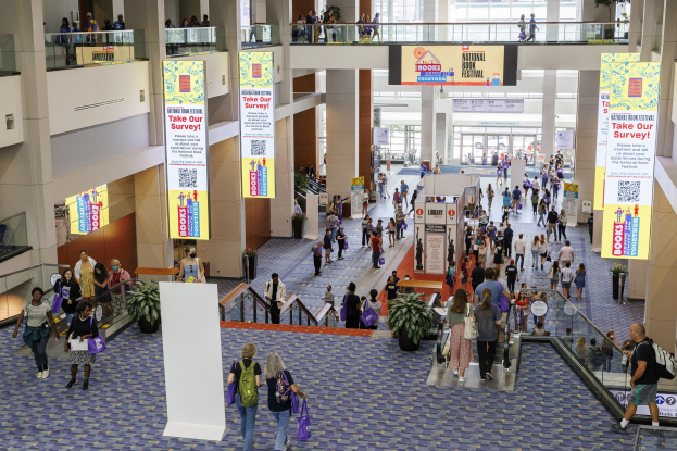 Eine große Menschenmenge bewegt sich durch ein großes Gebäude mit Bannern, Topfpflanzen, Rolltreppen, Geländern, Säulen, bedruckten Schildern, Glastüren und anderen Elementen, einige tragen Taschen, was auf den Besuch der Nationalen Buchmesse hinweist.