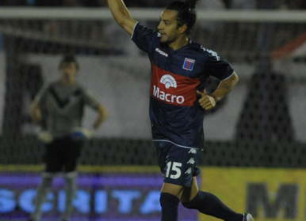 Ein Fußballspieler feiert nach einem Tor auf dem Boden, mit einer jubelnden Menge und einer Person im Hintergrund und einer Tafel mit Text dahinter, alles mit einem unscharfen, dynamischen Hintergrund.