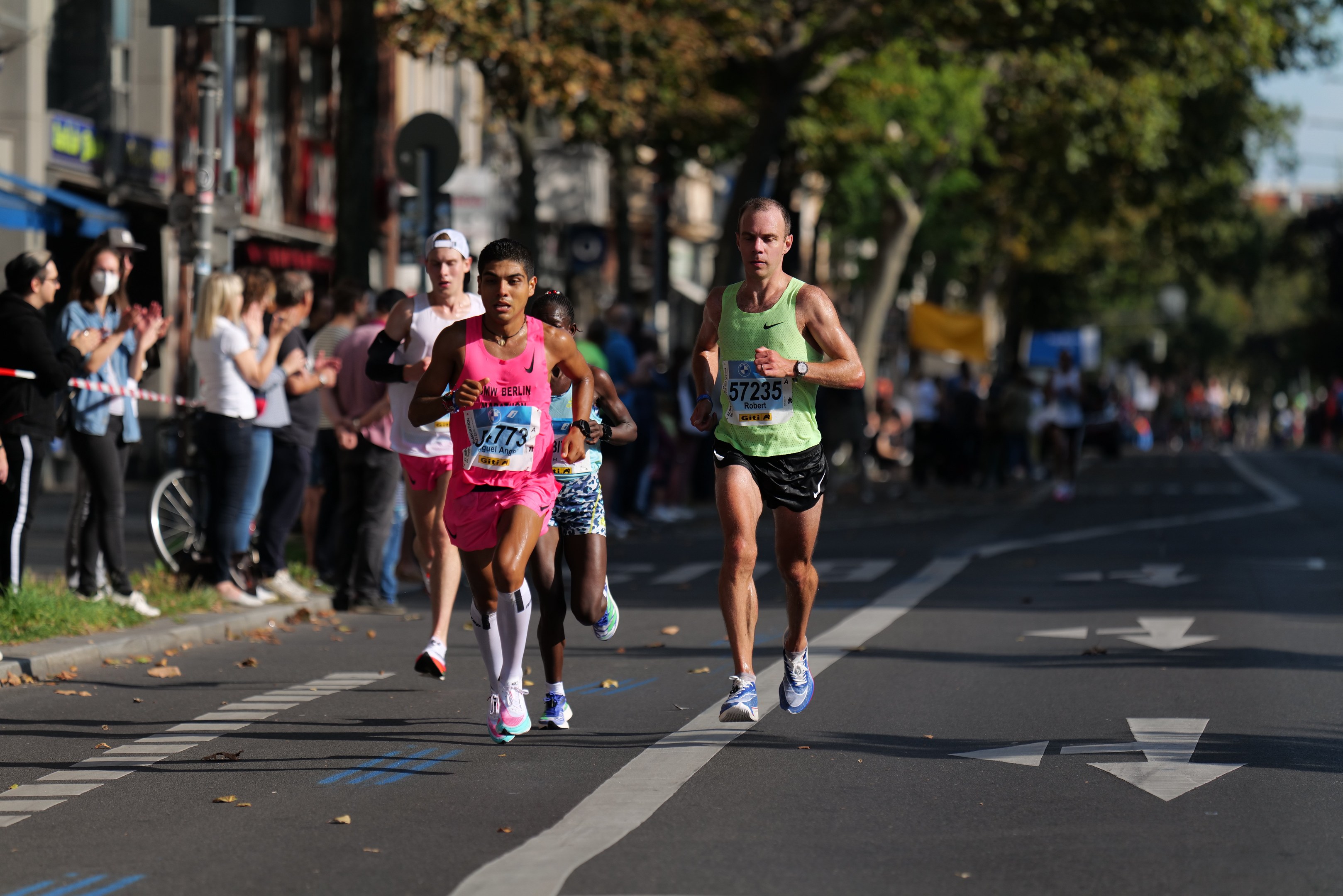 Eine Gruppe von Menschen, die bei einem Marathon auf einer Stadtstraße laufen, mit Zuschauern und unscharfen städtischen Elementen im Hintergrund.