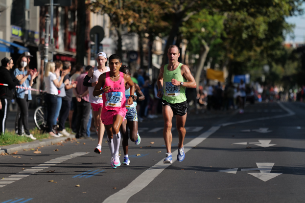 Eine Gruppe von Menschen, die bei einem Marathon auf einer Stadtstraße laufen, mit Zuschauern und unscharfen städtischen Elementen im Hintergrund.