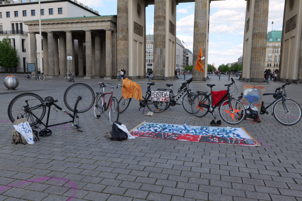Eine Gruppe von Fahrrädern, die vor dem Brandenburger Tor in Berlin, Deutschland, geparkt sind, mit Taschen und einem Banner in der Nähe, vor einer Kulisse aus Gebäuden, Bäumen, Pfählen, Flaggen und einem klaren blauen Himmel.