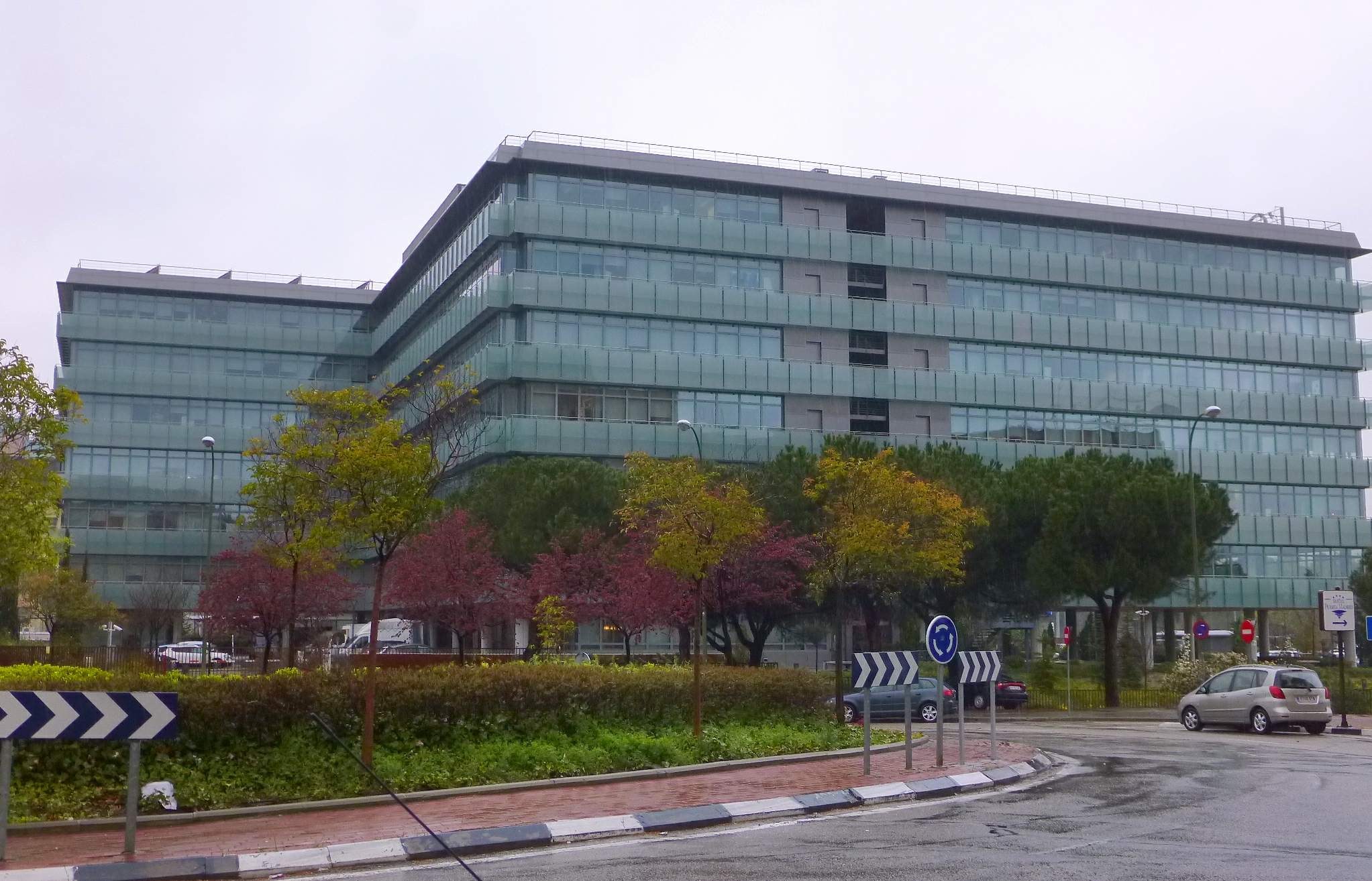 Großes modernes Gebäude mit zahlreichen Fenstern, der Hauptsitz der Europäischen Zentralbank, umgeben von Grünflächen, Straßeninfrastruktur und Fahrzeugen unter einem bewölkten Himmel.