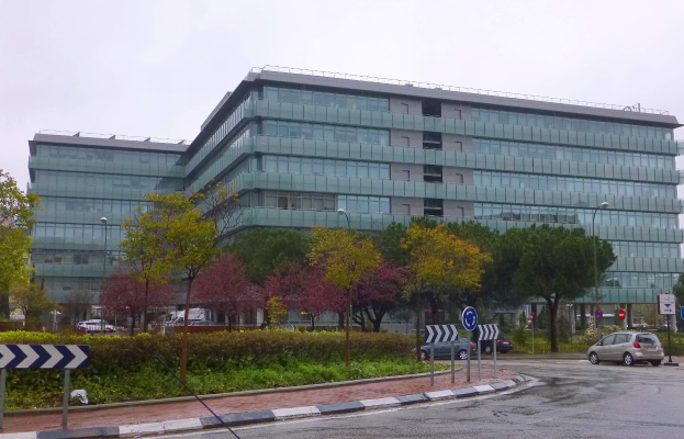 Großes modernes Gebäude mit zahlreichen Fenstern, der Hauptsitz der Europäischen Zentralbank, umgeben von Grünflächen, Straßeninfrastruktur und Fahrzeugen unter einem bewölkten Himmel.
