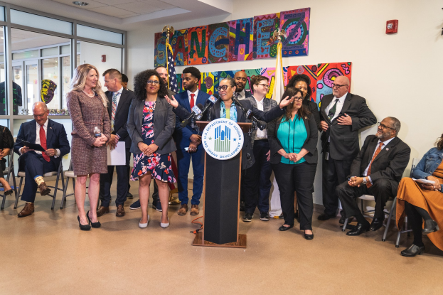 Eine Gruppe von Menschen steht vor einem Rednerpult mit einem Mikrofon und einer Tafel mit der Aufschrift "Sancho-Stadtrat tritt dem Stadtrat bei", während andere auf Stühlen sitzen oder in der Nähe stehen; Flaggen, ein Feuerlöscher und Deckenleuchten sind an der Rückwand zu sehen.