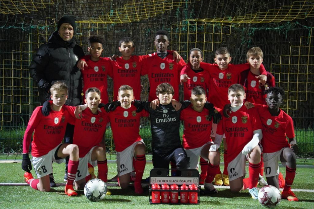 Eine Gruppe junger Jungs in roten T-Shirts und schwarzen Shorts posiert auf einem Fußballfeld mit verstreuten Bällen, Absperrungen und Bäumen im Hintergrund.