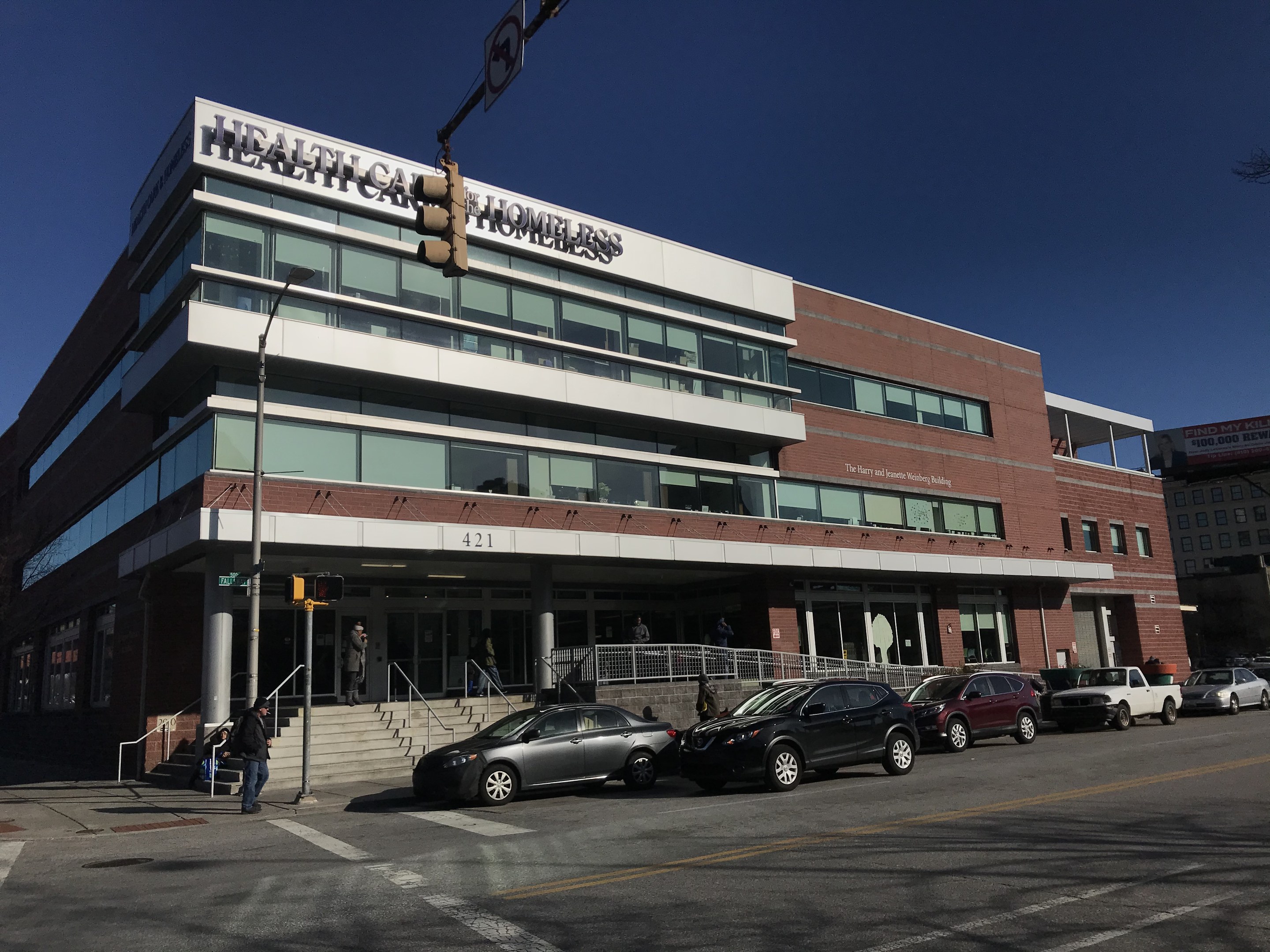 Großes Gebäude, das als New York City Health Care Homeless Center bezeichnet wird, mit geparkten Autos, Fußgängern, Straßenmöblierung und Bäumen unter einem klaren Himmel.