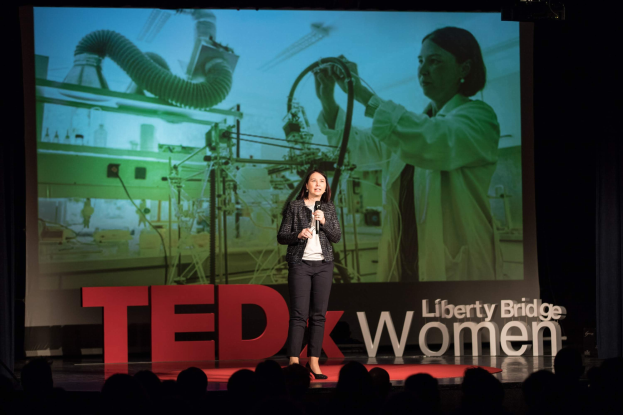 Frau auf einer Bühne mit Mikrofon, mit einem Display hinter ihr, das ein Bild einer Frau und Text zeigt.