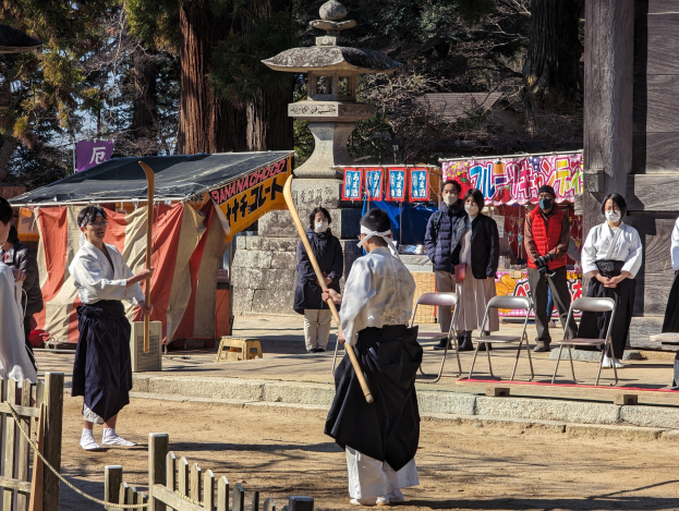 Eine Gruppe von Menschen in traditioneller Kleidung steht im Freien in Kyoto, einige tragen Masken und halten hölzerne Stäbe, während Stühle, Banner und ein Zelt gegen einen klaren blauen Himmel sichtbar sind.