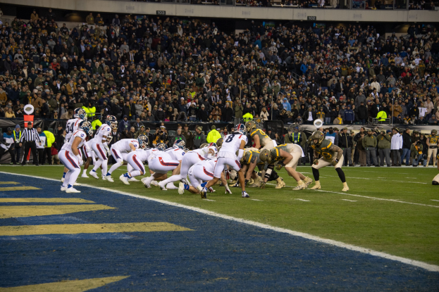 Ein Fußballspiel im Gange mit Spielern auf dem Boden in Sportbekleidung und Helmen, aus der Perspektive eines Stadionplatzes mit Zuschauern und Werbetafeln im Hintergrund.