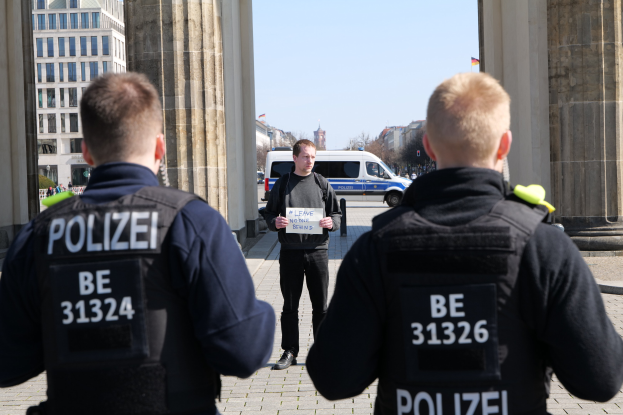 Three police officers stand in front of a building, with a man in the middle holding a paper, flanked by pillars and a busy urban background.