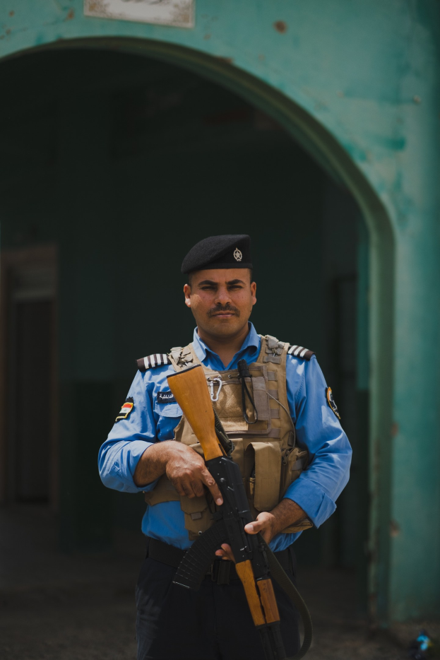 Polizeibeamter in Uniform mit Gewehr vor einem Gebäude mit einem Torbogen und einer Tafel an der Wand.