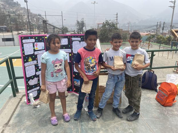 Gruppe von Kindern auf einem Basketballfeld mit Papiertüten in den Händen, mit Tüten und Gegenständen rechts und einer Tafel mit Text links, vor einer Außenkulisse mit Zaun, Pfosten, Straßenlaternen, Strommasten, Bäumen, Gebäuden, Hügeln und Himmel.