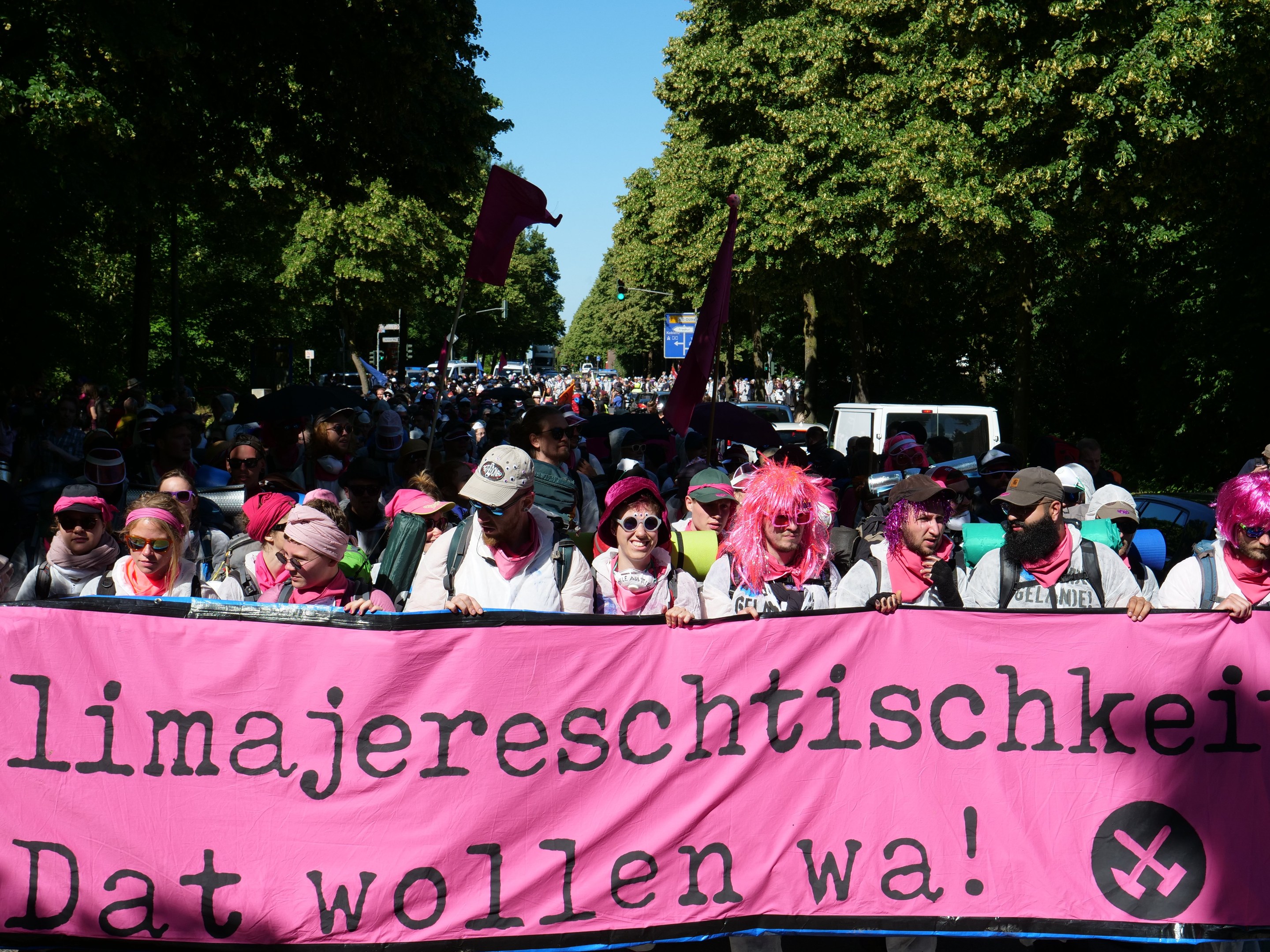 Eine Gruppe von Menschen marschiert die Straße entlang und hält ein pinkes Banner mit der Aufschrift "Women's March in Berlin", während einige eine Mütze und eine Schutzbrille tragen und andere Fahnen halten, vor dem Hintergrund von Bäumen, Fahrzeugen und Himmel.