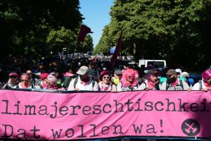 Eine Gruppe von Menschen marschiert die Straße entlang und hält ein pinkes Banner mit der Aufschrift "Women's March in Berlin", während einige eine Mütze und eine Schutzbrille tragen und andere Fahnen halten, vor dem Hintergrund von Bäumen, Fahrzeugen und Himmel.