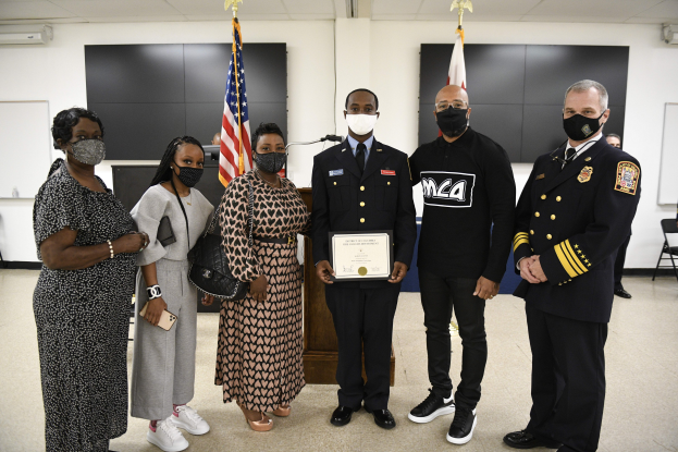 Eine Gruppe von Menschen in Masken steht zusammen vor Flaggen, hält ein Zertifikat hoch, wobei die zentrale Person es deutlich zur Schau stellt, in einer Umgebung, die wie eine Abschlussfeier mit Stühlen, Tischen und anderen Gegenständen im Hintergrund aussieht.