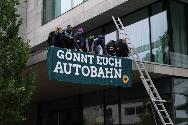 Gruppe von Menschen auf einem Dach, die ein Protestplakat halten, mit einem Baum links und Glasfenstern im Hintergrund.
